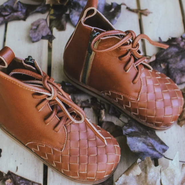 Pair of brown leather shoes with woven pattern on a wooden surface with leaves.