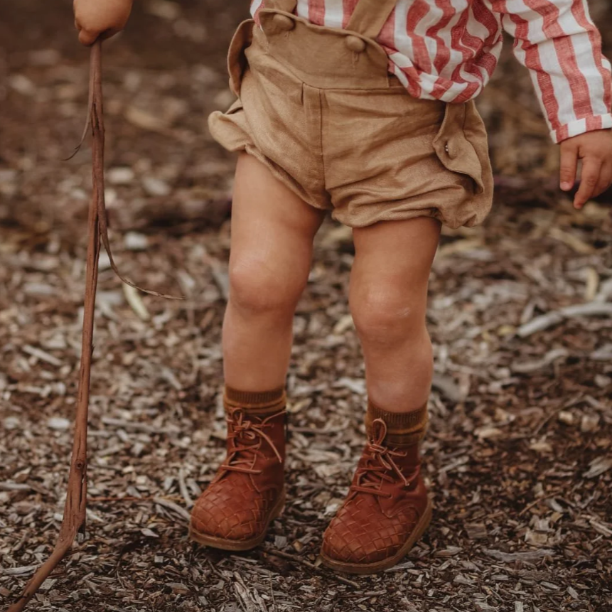 Child wearing a red and white striped shirt, beige shorts, and brown boots standing on a mulch-covered ground.