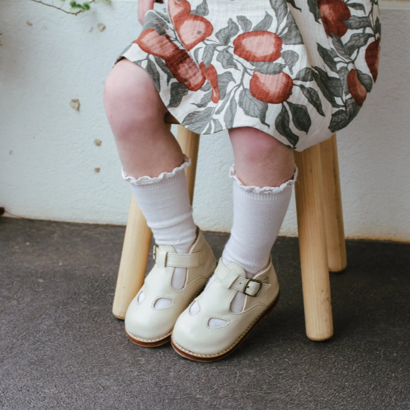 Child wearing white shoes and socks sitting on a wooden stool with a floral dress.