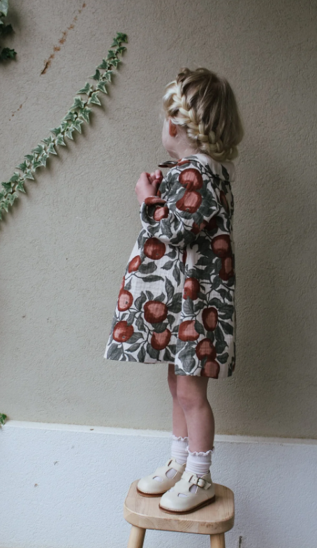 Child wearing a patterned dress standing on a small wooden stool against a plain wall.