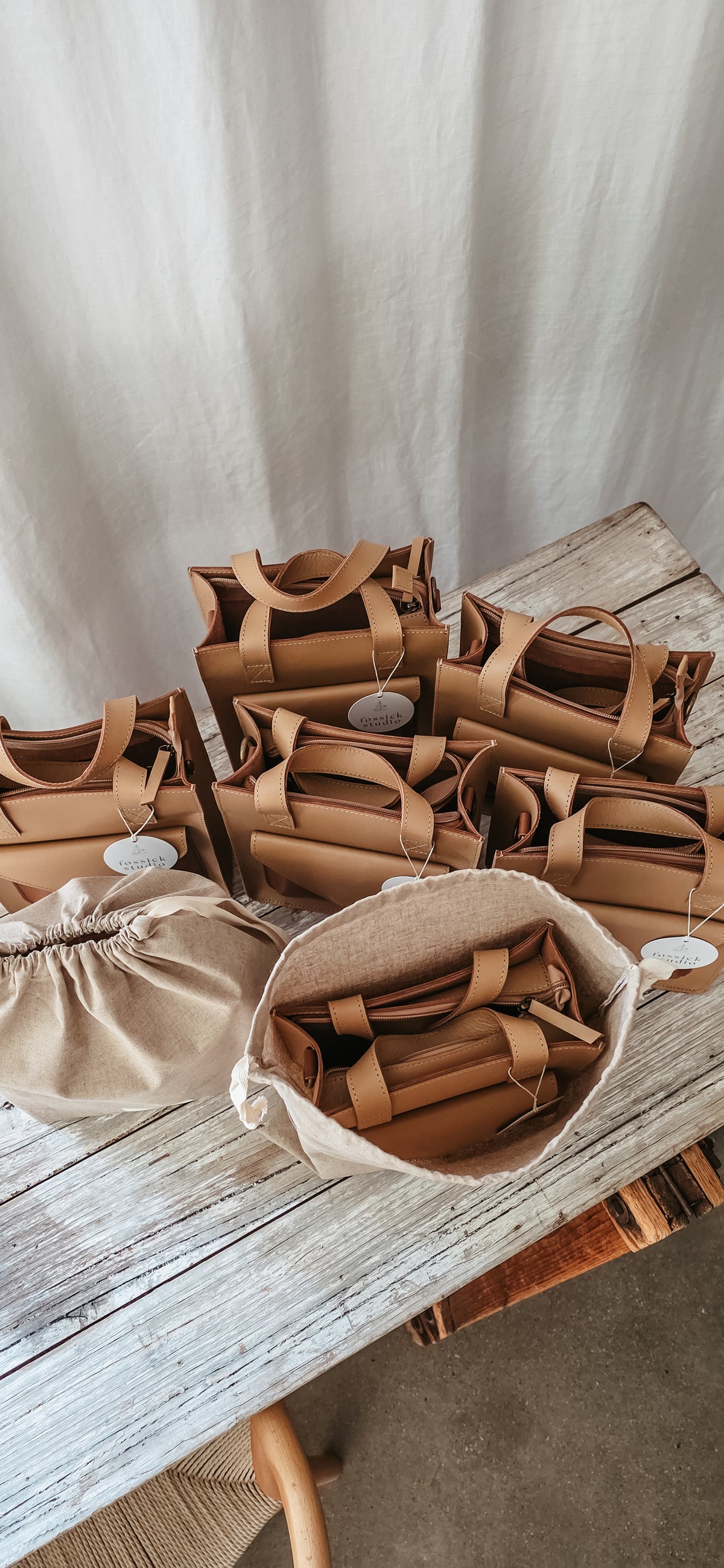 A styled arrangement of multiple tan leather and canvas tote bags on a rustic wooden table against a neutral draped fabric backdrop.