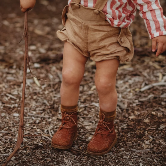 Child wearing a red and white striped shirt, beige shorts, and brown boots standing on a mulch-covered ground.