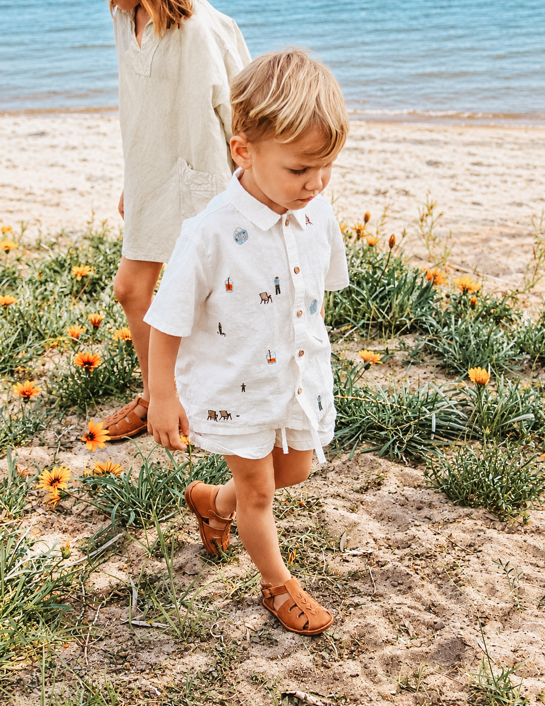 A young child walking on a sandy beach path with flowers, wearing a white short-sleeve button-up shirt and matching shorts with small embroidered details, paired with brown sandals. Another child in a cream dress stands behind in the background near the ocean.