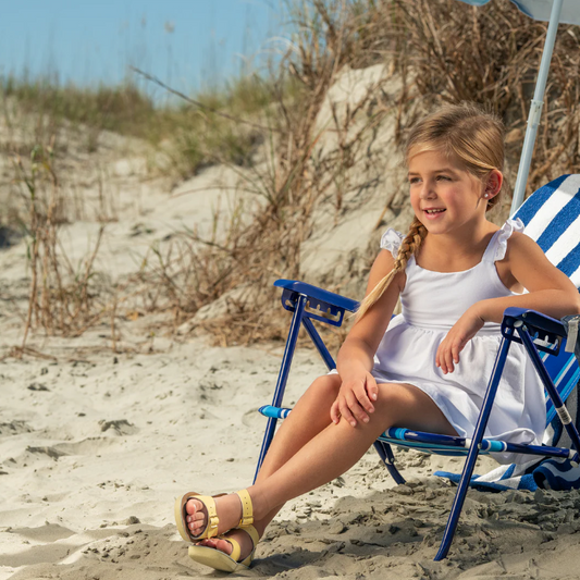 Young girl in a white dress sitting on a blue and white striped beach chair on a sandy beach.